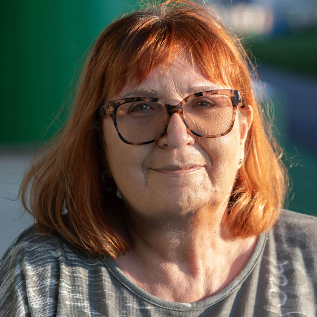 Heidi hat rote Haaren und blaugraue Augen und trägt eine braun gemusterte Brille. Sie lächelt in die Kamera und sanftes Sonnenlicht fällt von rechts auf ihr Gesicht.