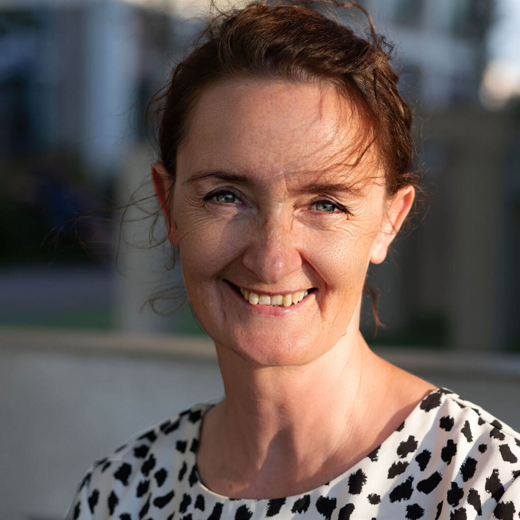 Katarzyna hat rotbraune Haare und blaue Augen. Sie trägt ein schwarz gepunktetes Oberteil und lächelt in die Kamera, während sanftes Sonnenlicht von rechts ihr Gesicht beleuchtet.