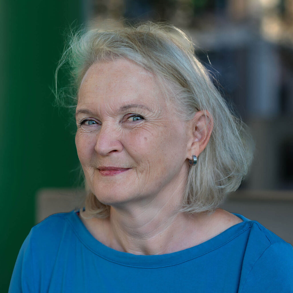 Michaela hat graue Haare, blaue Augen und trägt ein blaues Oberteil. Sie hat den Kopf leicht von der Kamera abgewendet und lächelt.