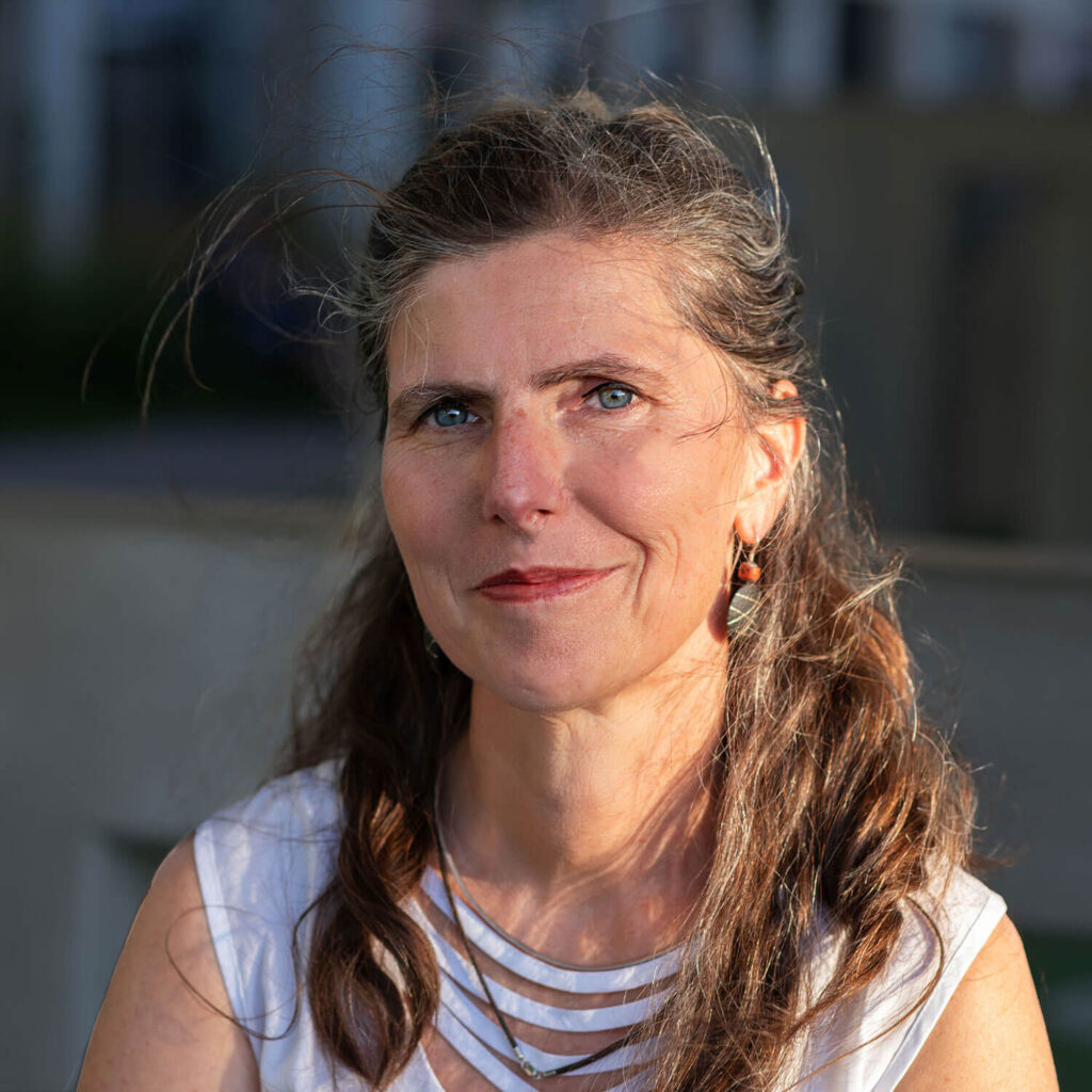 Sonja hat grau-braune Haare, blaue Augen, trägt ein weißes Oberteil und dunkelroten Lippenstift. Sie lächelt, während Sonnenlicht von rechts auf ihr Gesicht fällt.