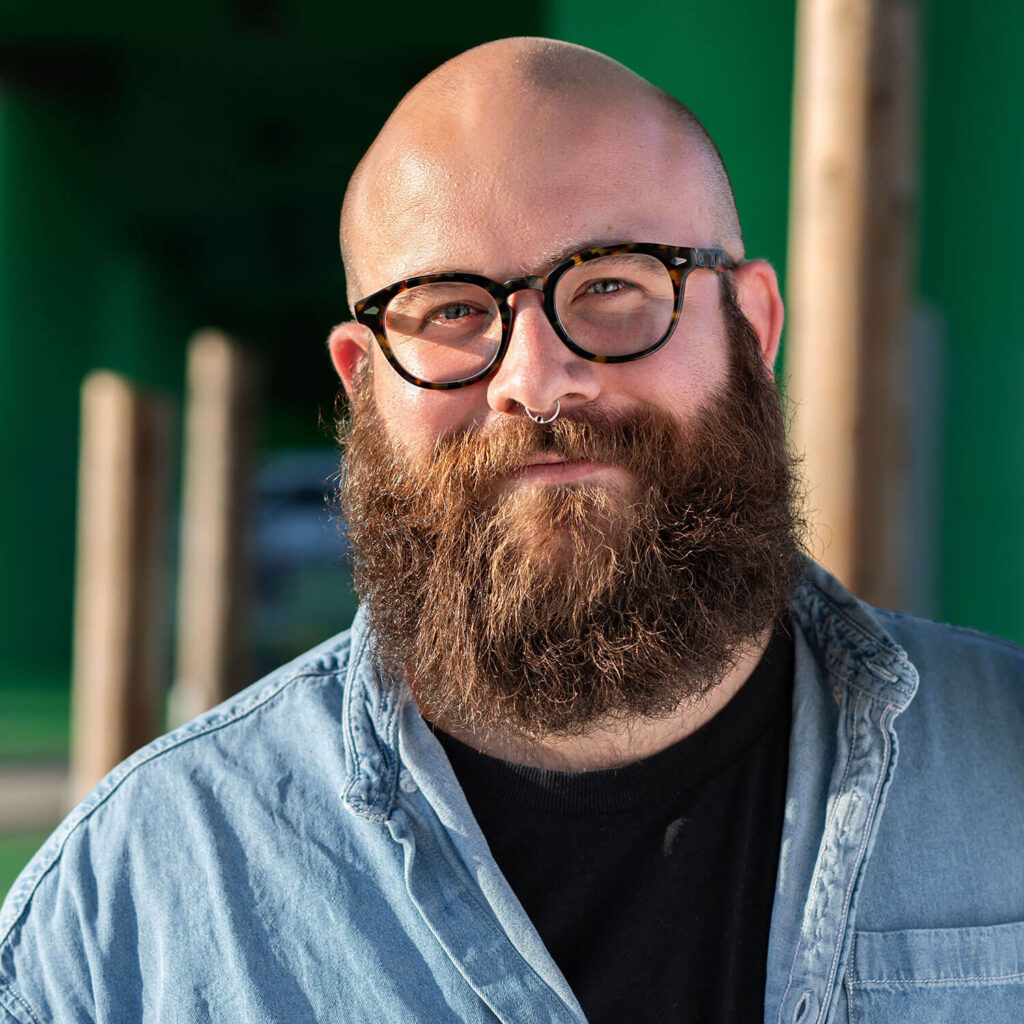 Timo hat eine Glatze, blaue Augen und einen braunen Vollbart. Timo trägt eine schwarz-braune Brille, hat ein Nasenpiercing und trägt ein Jeanshemd. Timo lächelt in die Kamera, während Sonnenlicht von links auf Timos Gesicht fällt.